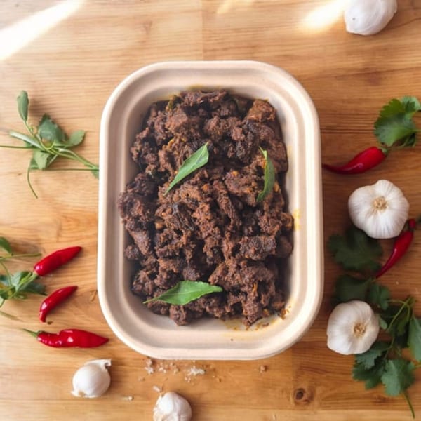Indian beef fry in a container, surrounded by red chilies, garlic, and cilantro on a wooden table.