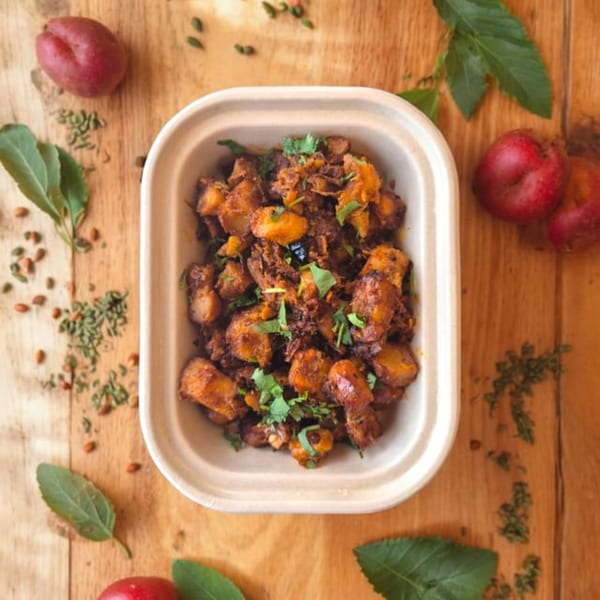 Indian potato dish with cilantro in a takeout container, surrounded by spices and red plums.