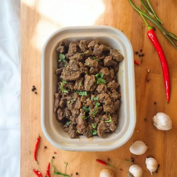 Indian beef fry in a takeaway container, garnished with cilantro, on a wooden board with chilies and garlic.