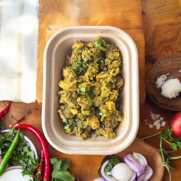 Top-down view of Kolapasi Indian Canteen's green curry in a takeout container, surrounded by fresh ingredients.