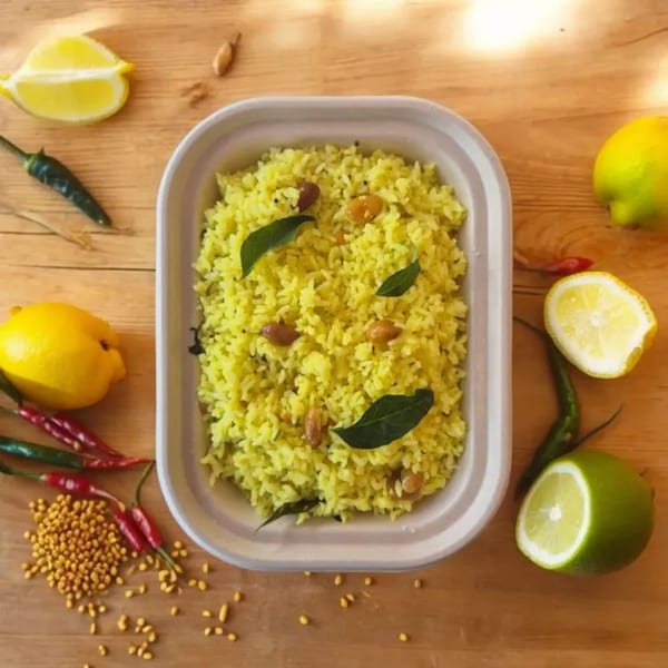 Yellow lemon rice with peanuts and curry leaves in a container, surrounded by lemons and chilies.